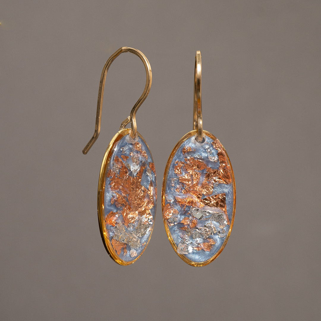 A pair of gold-tone dangle earrings with oval clear resin pendants containing embedded copper and light blue glitter flakes, shown against a solid gray background.
