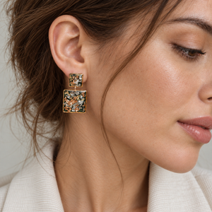 A close-up of a woman wearing gold square drop earrings with colorful floral enamel detailing in orange, green, and blue.