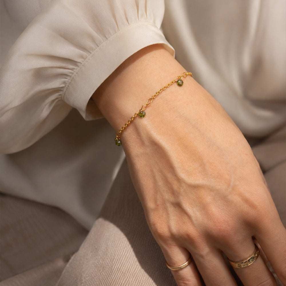 A delicate gold chain bracelet with small green faceted bead charms, displayed on a clean off-white background.