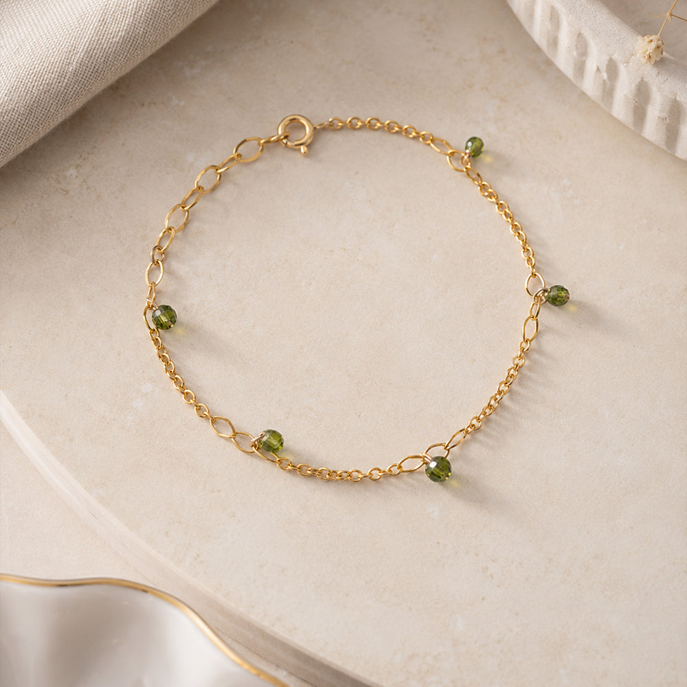 A delicate gold chain bracelet with small green faceted bead charms, displayed on a clean off-white background.