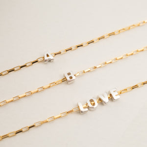 Three gold chain bracelets with silver letter charms spelling out 'A', 'B', and 'LOVE' on a light beige background.