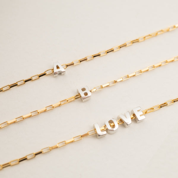 Three gold chain bracelets with silver letter charms spelling out 'A', 'B', and 'LOVE' on a light beige background.