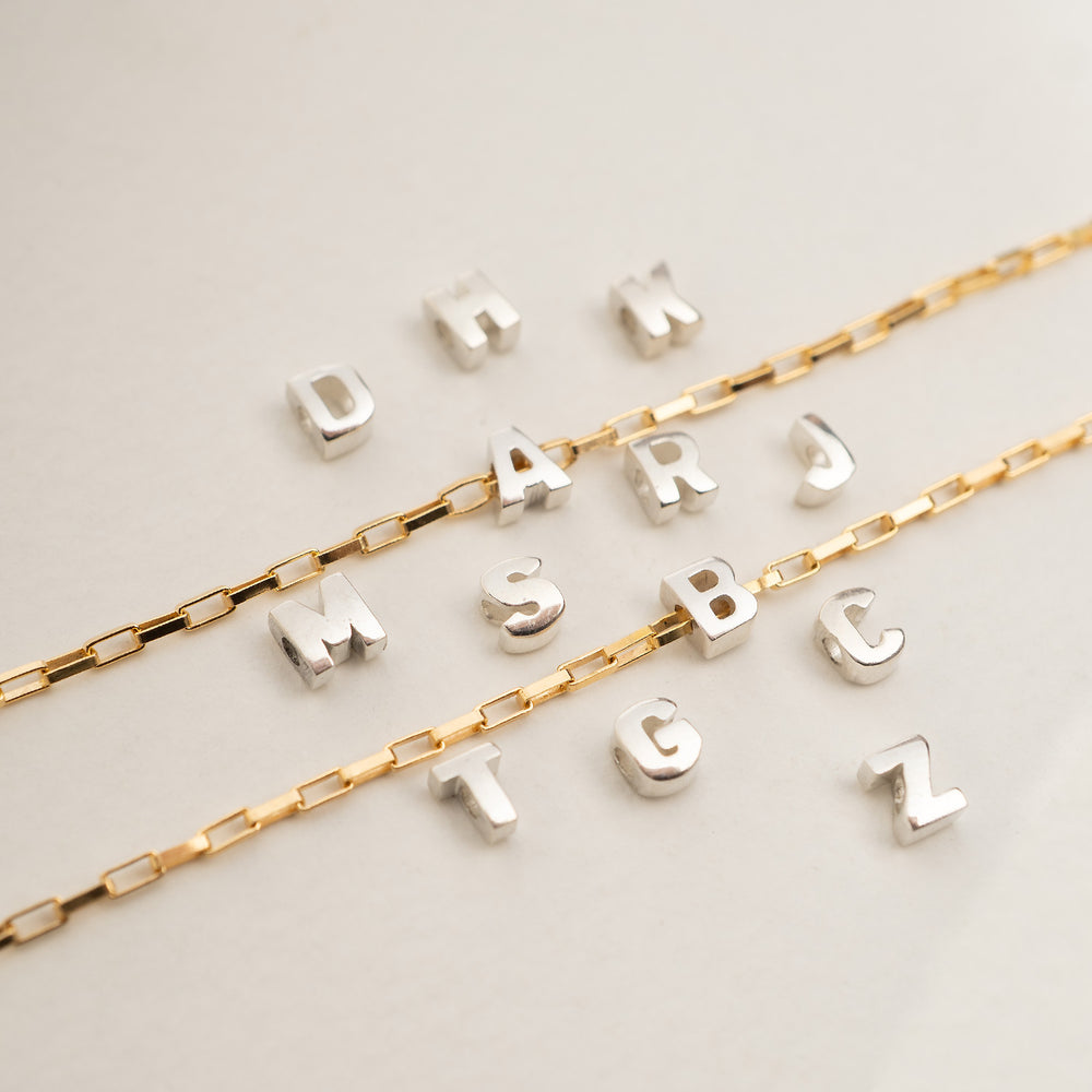 Two gold-tone chain necklaces laid on a light neutral background, each paired with a row of silver-tone initial charms spelling out names.