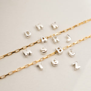 Two gold-tone chain necklaces laid on a light neutral background, each paired with a row of silver-tone initial charms spelling out names.