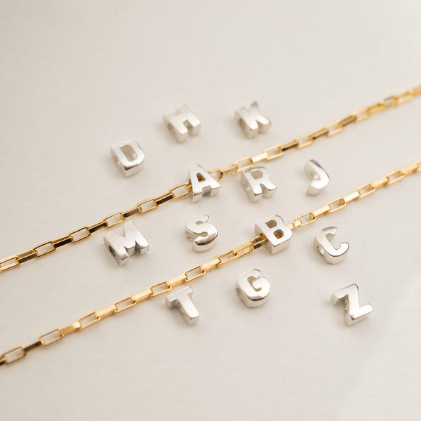 Two gold-tone chain necklaces laid on a light neutral background, each paired with a row of silver-tone initial charms spelling out names.