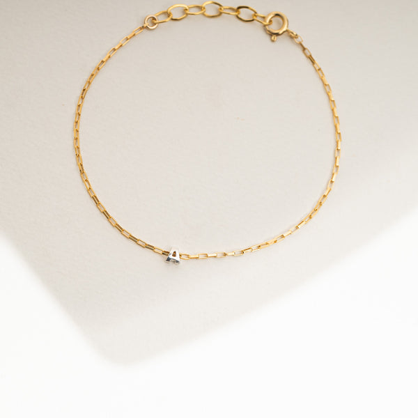 A delicate gold chain bracelet with a small silver bow charm, displayed on a light beige surface with a soft shadow.