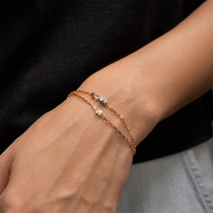 A close-up of a person's wrist wearing two delicate bracelets: one gold chain bracelet with a small silver letter 'A' charm and one gold chain bracelet with a small silver letter 'M' charm, worn over a black top.