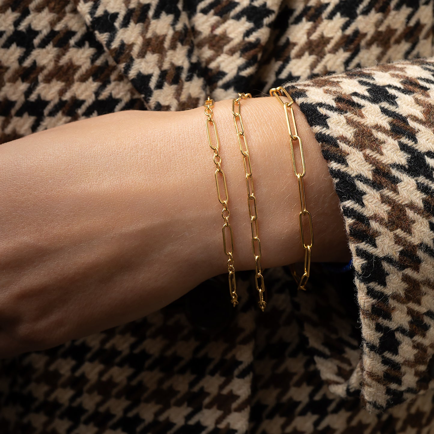 Gold chain bracelets on a wrist with a houndstooth patterned fabric background