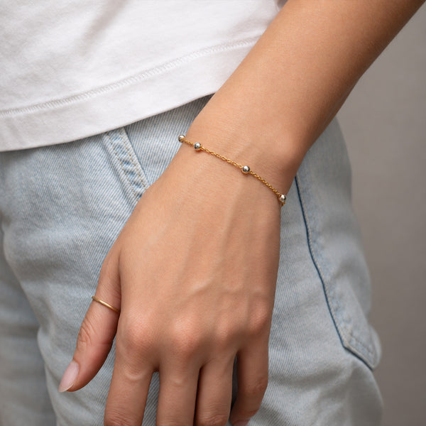 A delicate gold chain bracelet with small silver bead accents worn on a person's wrist, paired with a simple gold ring on the finger, shown against a light gray denim background.