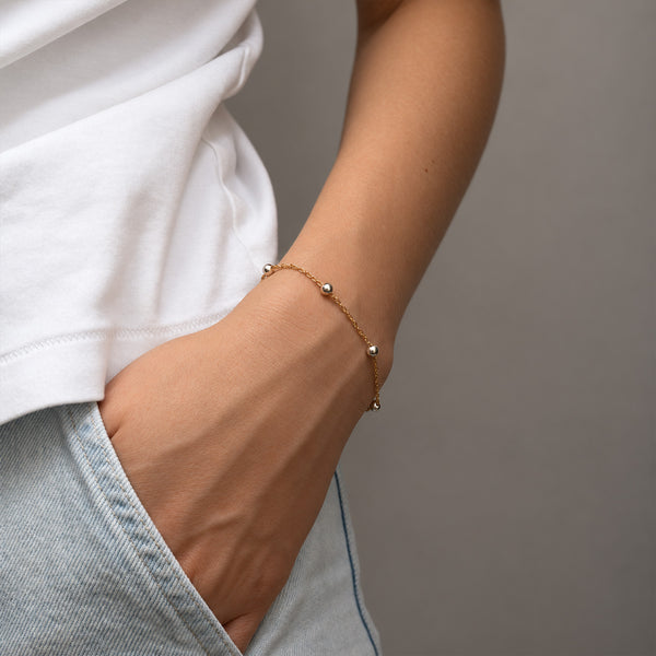 A delicate gold chain bracelet with small silver bead accents worn on a person's wrist, shown against a neutral gray background with a white t-shirt and light denim shorts.