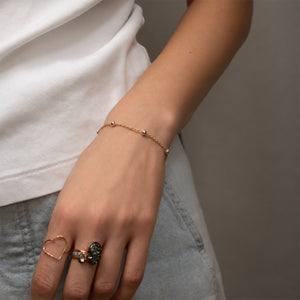 A close-up of a person's hand and wrist wearing a delicate gold chain bracelet with small silver bead accents, along with rings, against a neutral gray background.