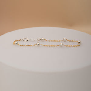 A delicate gold chain bracelet with five polished silver bead accents, displayed on a white cylindrical pedestal against a warm beige background.