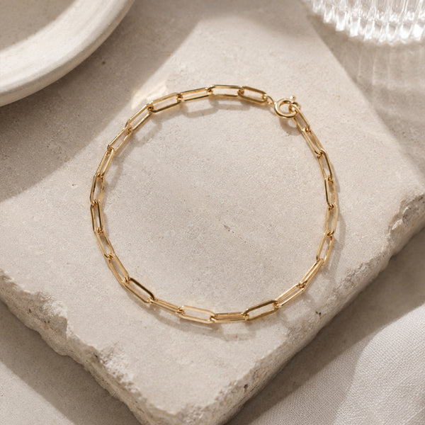 A gold paperclip chain bracelet displayed on a textured light beige stone block with soft, warm lighting.