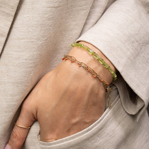 A close-up of a person's hand wearing two delicate bracelets: one green beaded bracelet and one gold chain bracelet with small orange-red bead accents, set against a beige linen garment.
