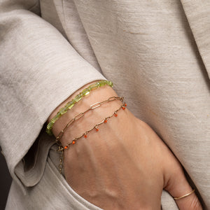 A close-up of a person's hand and wrist wearing three delicate bracelets: a lime green beaded bracelet, a gold paperclip bracelet, and a gold chain with small orange beads, all styled with a light beige linen jacket.