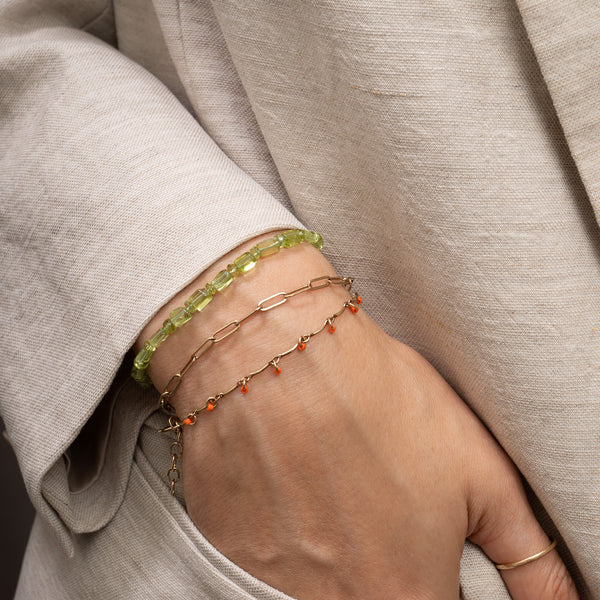 A close-up of a person's hand and wrist wearing three delicate bracelets: a lime green beaded bracelet, a gold paperclip bracelet, and a gold chain with small orange beads, all styled with a light beige linen jacket.