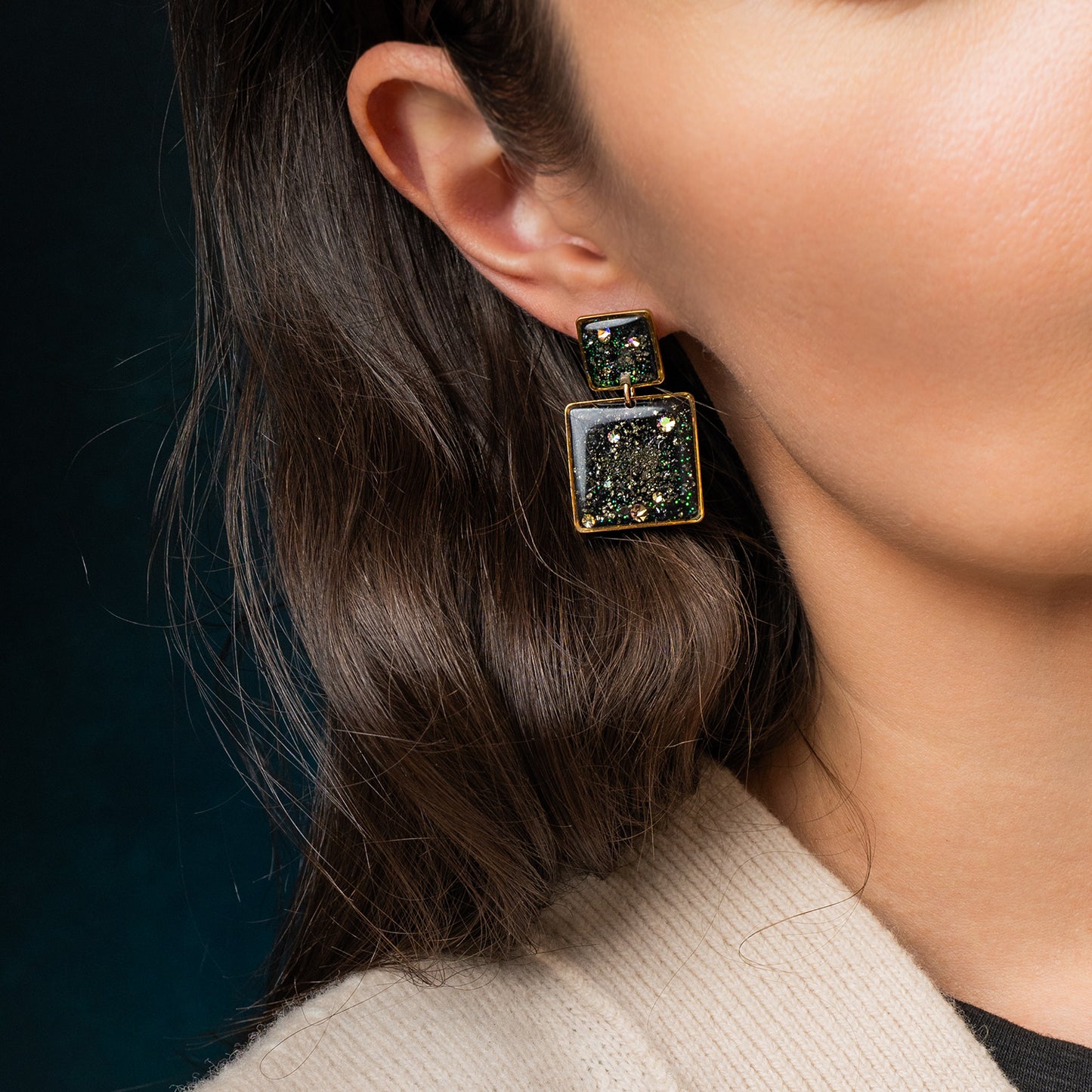 Close-up of a person wearing a pair of square earrings with green gemstones against a dark background.
