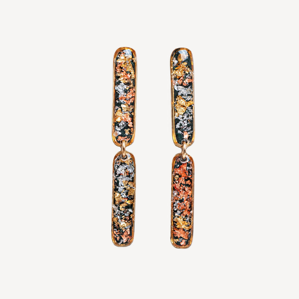 A pair of gold-toned drop earrings with elongated rectangular charms featuring a multicolor metallic glitter pattern of orange, gold, silver, and dark green, set against a plain off-white background.