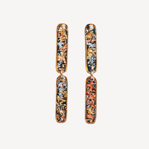A pair of gold-toned drop earrings with elongated rectangular charms featuring a multicolor metallic glitter pattern of orange, gold, silver, and dark green, set against a plain off-white background.