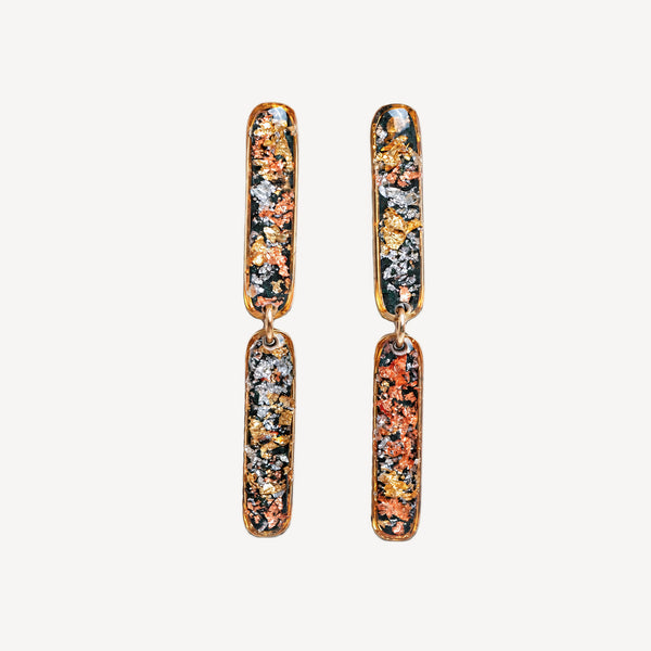 A pair of gold-toned drop earrings with elongated rectangular charms featuring a multicolor metallic glitter pattern of orange, gold, silver, and dark green, set against a plain off-white background.