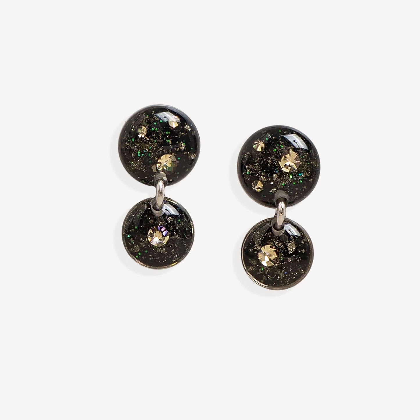 A pair of double-drop statement earrings featuring round black resin pendants filled with gold glitter and iridescent flecks, suspended from silver-toned metal rings on a light gray background.