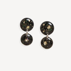 A pair of double-drop statement earrings featuring round black resin pendants filled with gold glitter and iridescent flecks, suspended from silver-toned metal rings on a light gray background.