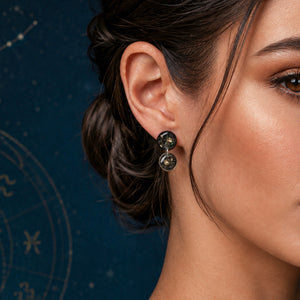 A close-up of a woman's ear and side profile wearing a pair of dark, textured drop earrings with small circular gold-toned details, set against a dark blue background with subtle zodiac constellation graphics.