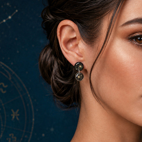 A close-up of a woman's ear and side profile wearing a pair of dark, textured drop earrings with small circular gold-toned details, set against a dark blue background with subtle zodiac constellation graphics.