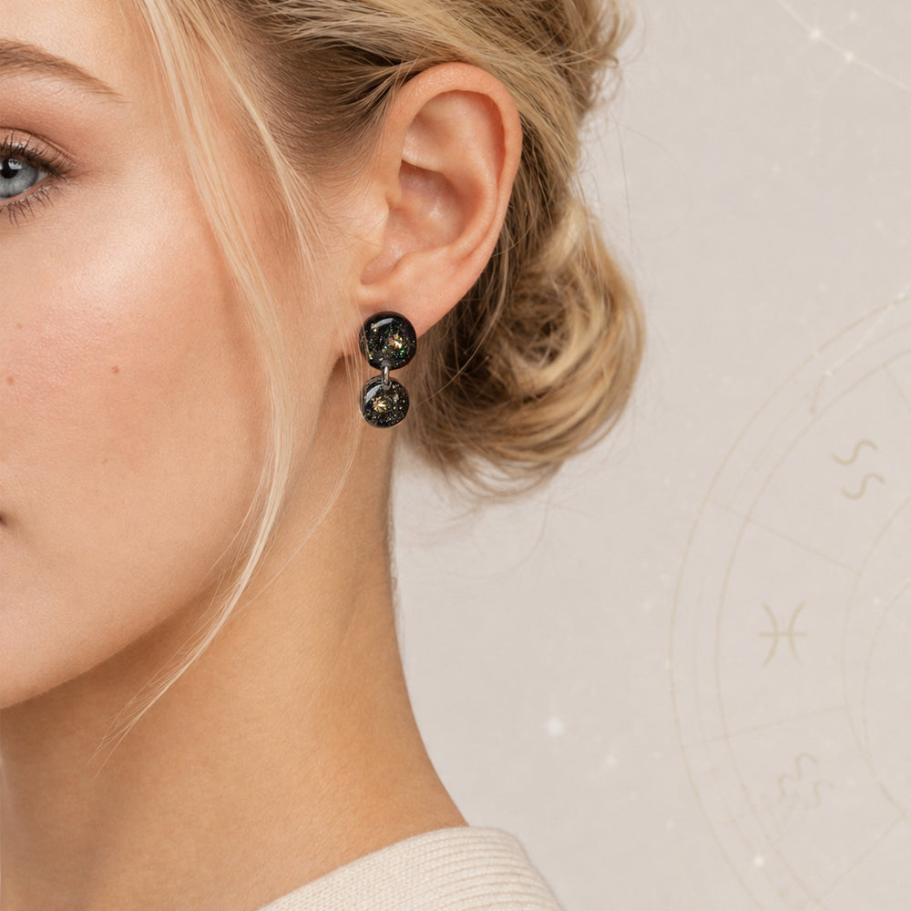A pair of double-drop statement earrings featuring round black resin pendants filled with gold glitter and iridescent flecks, suspended from silver-toned metal rings on a light gray background.