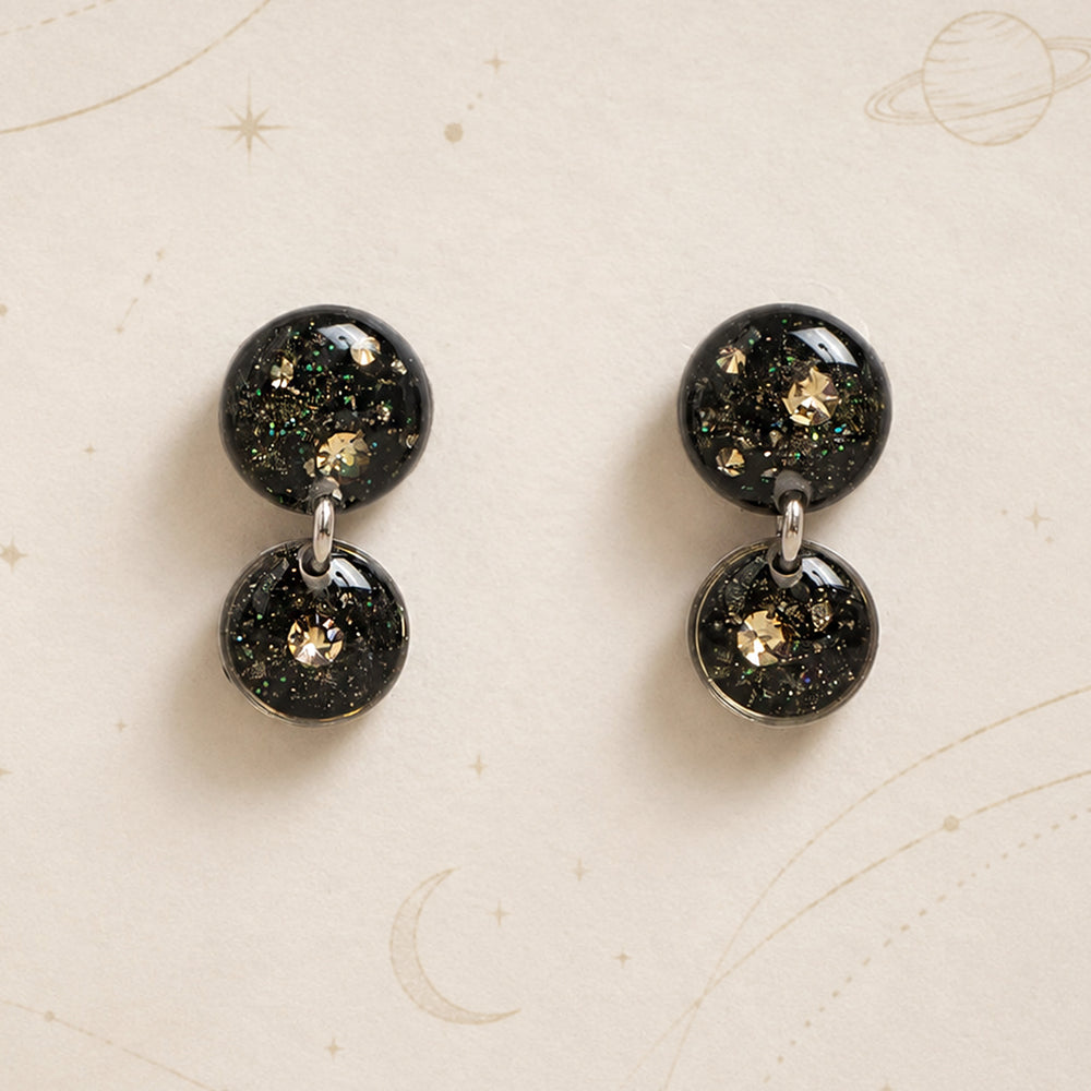 A pair of double-drop statement earrings featuring round black resin pendants filled with gold glitter and iridescent flecks, suspended from silver-toned metal rings on a light gray background.