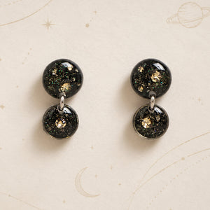 A pair of double-stud dangle earrings with glossy black spherical studs filled with gold glitter and iridescent flecks, displayed on a cream-colored background with celestial line drawings.