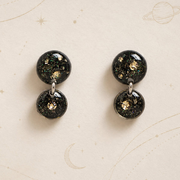 A pair of double-stud dangle earrings with glossy black spherical studs filled with gold glitter and iridescent flecks, displayed on a cream-colored background with celestial line drawings.