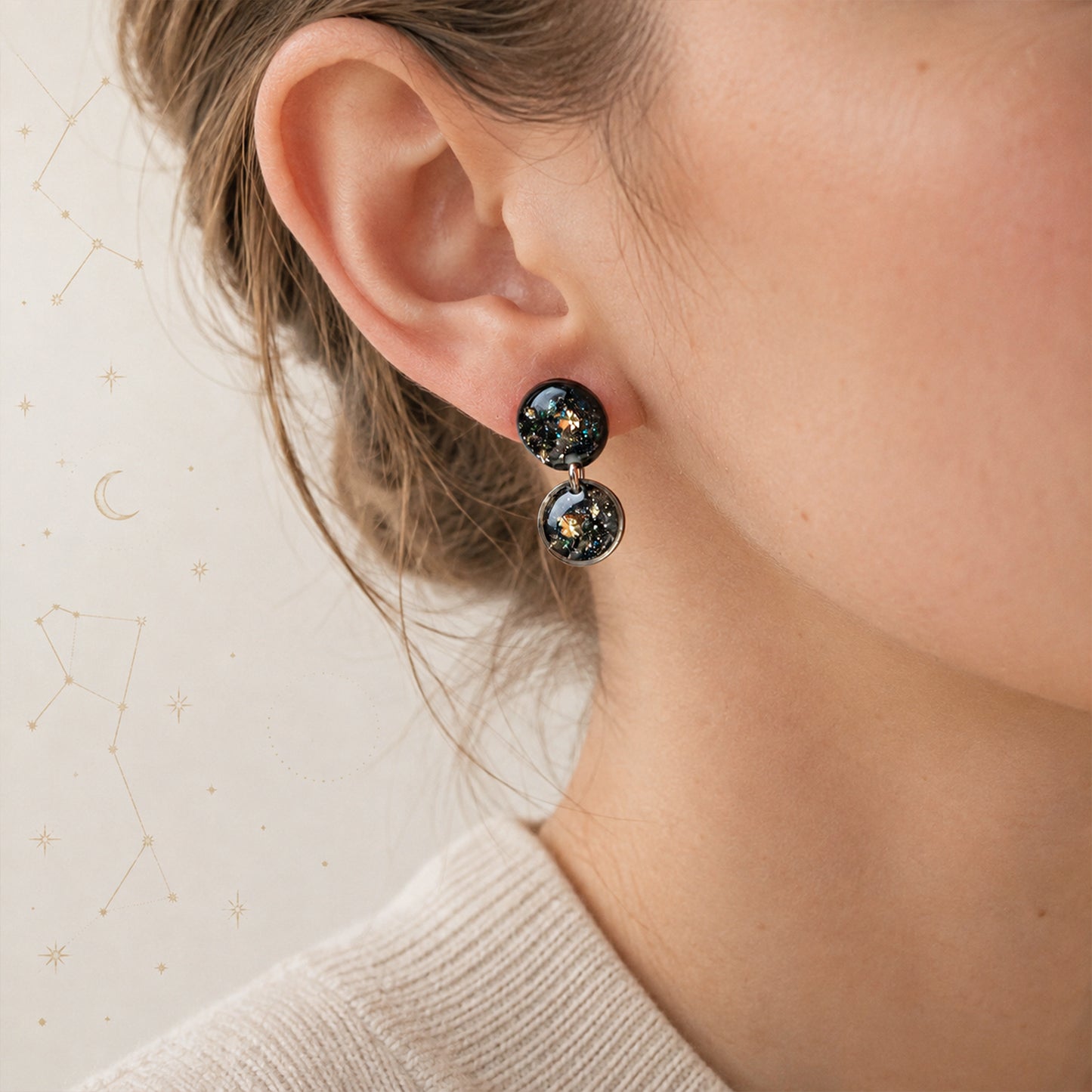 A close-up of a person's ear wearing a pair of dangling round earrings with a black glittery space design featuring small gold stars.