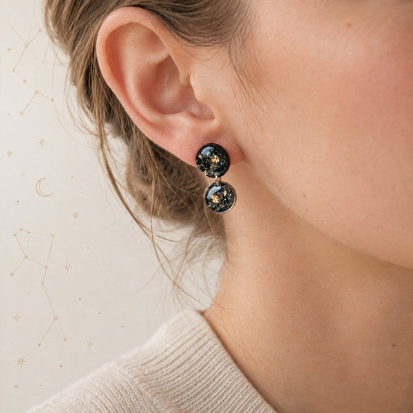A close-up of a person's ear wearing a pair of dangling round earrings with a black glittery space design featuring small gold stars.