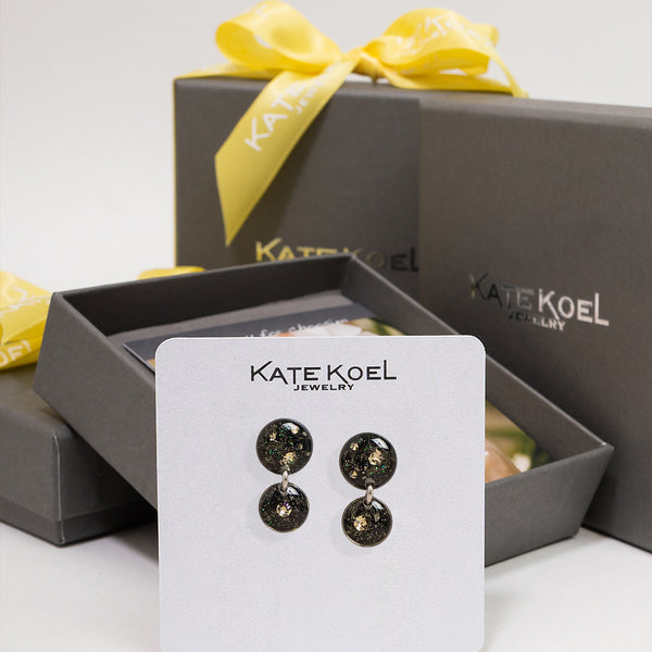 Kate Koel Jewelry double drop earrings with round black and gold glitter drops, displayed on a white branded card in front of open gray gift boxes with yellow ribbon.
