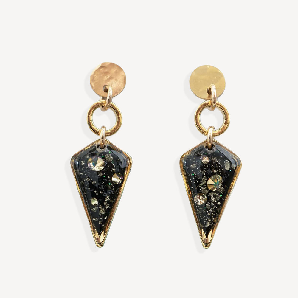 A pair of gold-tone dangle earrings with round hammered studs, circular jump rings, and pointed triangular resin pendants filled with black glitter and iridescent flecks, shown on a light gray background.