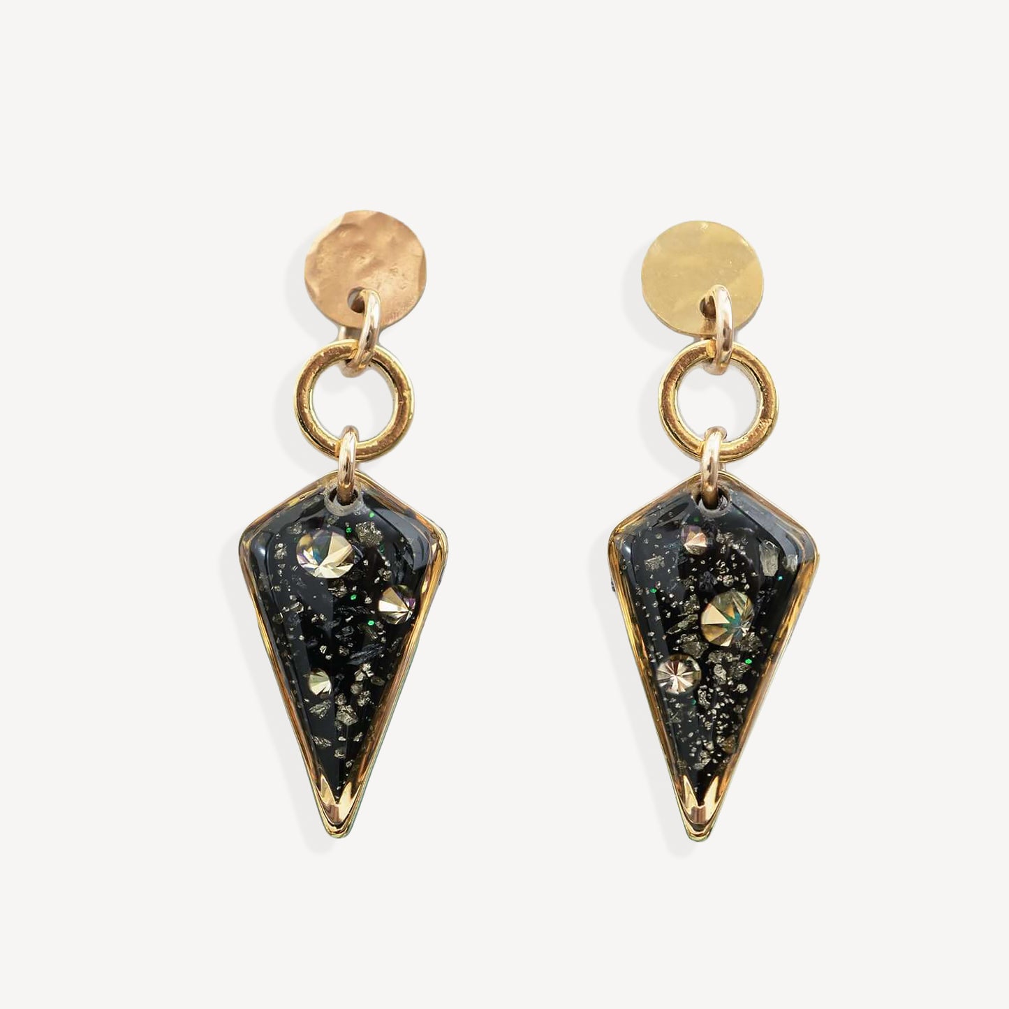 A pair of gold-tone dangle earrings with round hammered studs, circular jump rings, and pointed triangular resin pendants filled with black glitter and iridescent flecks, shown on a light gray background.