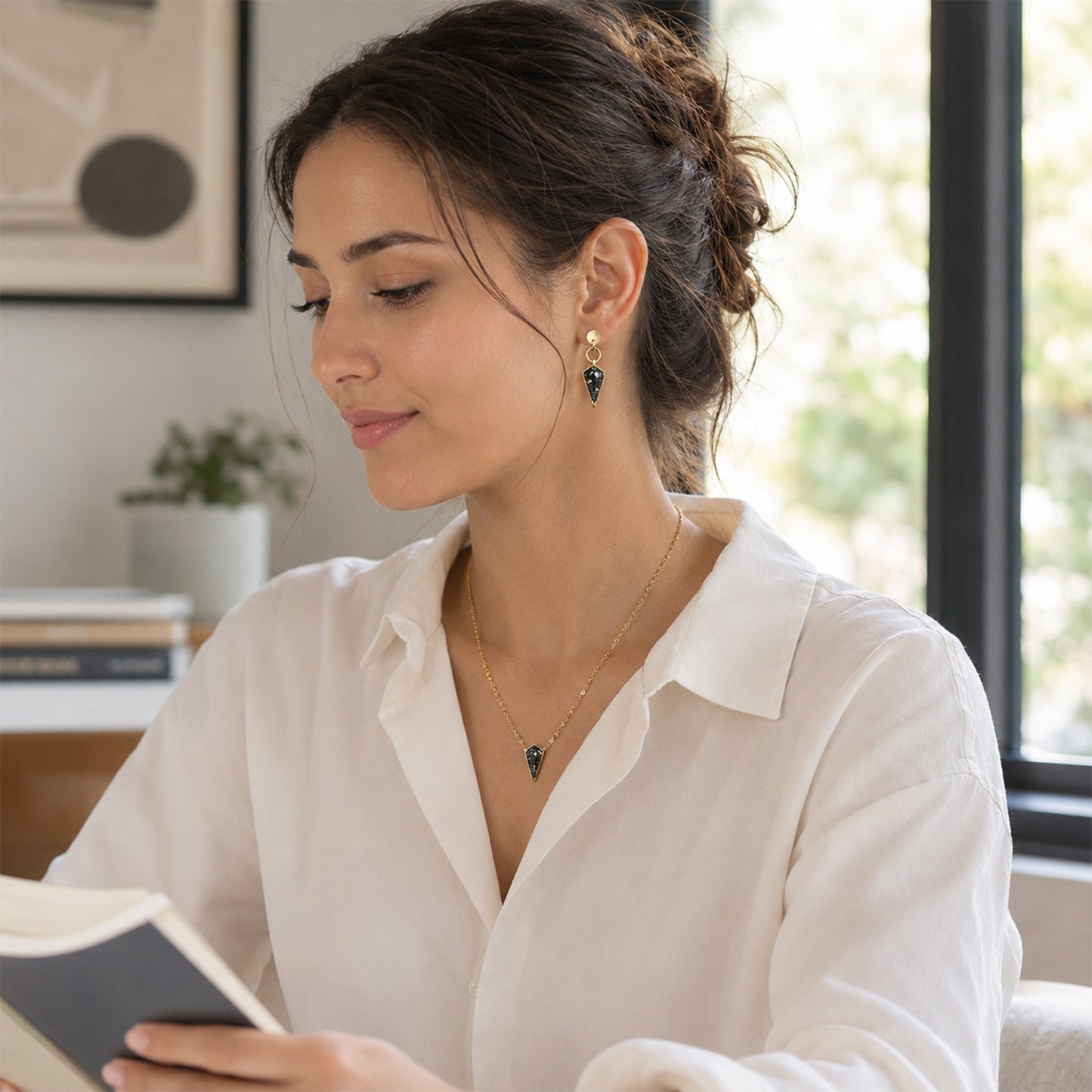 A woman in a white collared shirt wearing a delicate gold necklace and matching drop earrings with dark blue stones, sitting indoors and reading a book.