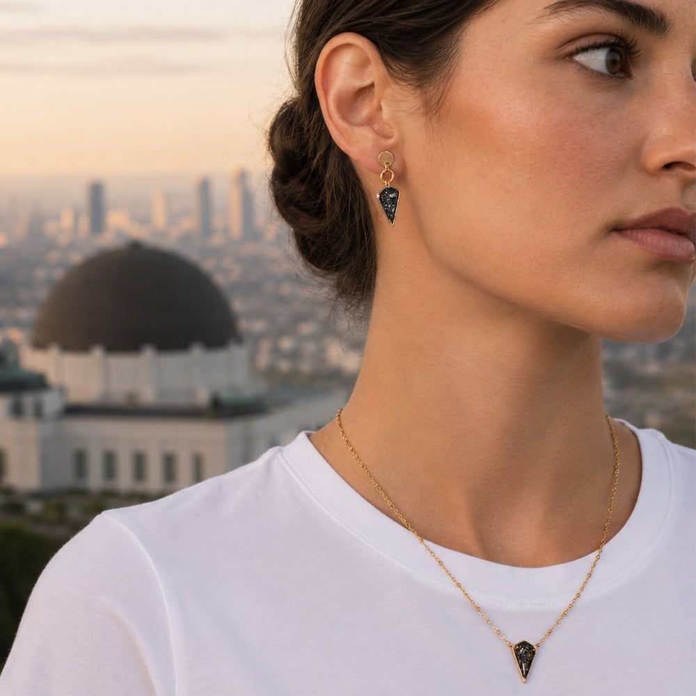 A pair of gold-tone dangle earrings with round hammered studs, circular jump rings, and pointed triangular resin pendants filled with black glitter and iridescent flecks, shown on a light gray background.