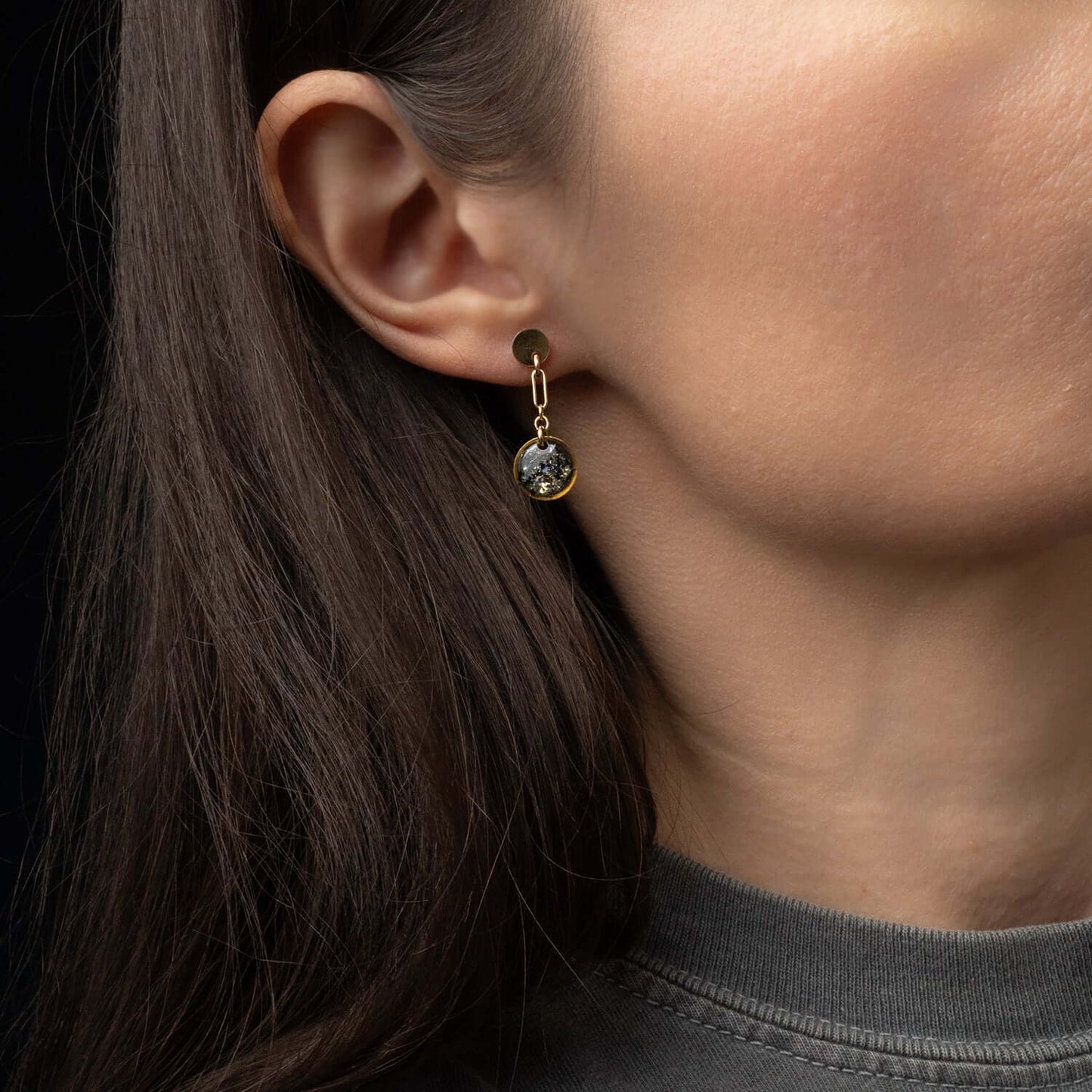 Close-up of an earring worn by a person with dark hair and a gray shirt on a black background