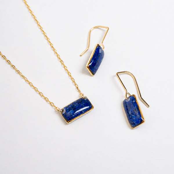 A gold jewelry set featuring a necklace and matching drop earrings, both with rectangular blue lapis lazuli stones in bezel settings, displayed on a clean white background.