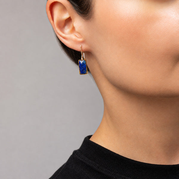 A close-up of a person's ear and neck wearing a small rectangular drop earring with a deep blue stone in a gold-toned setting.
