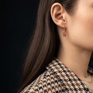 A close-up side profile of a woman wearing a small gold hoop earring with a rectangular amber stone, against a dark background.