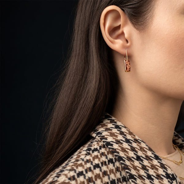 A close-up side profile of a woman wearing a small gold hoop earring with a rectangular amber stone, against a dark background.