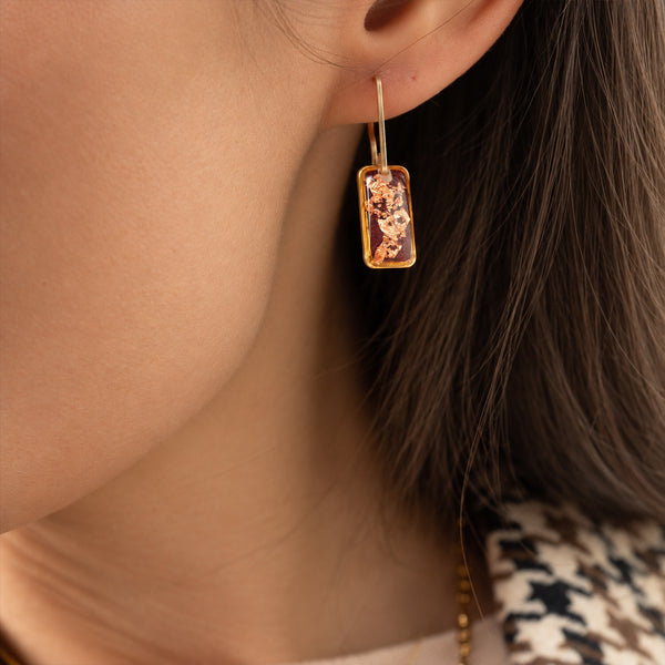 A close-up of a model wearing a gold rectangular drop earring with a textured, organic amber-orange inlay.