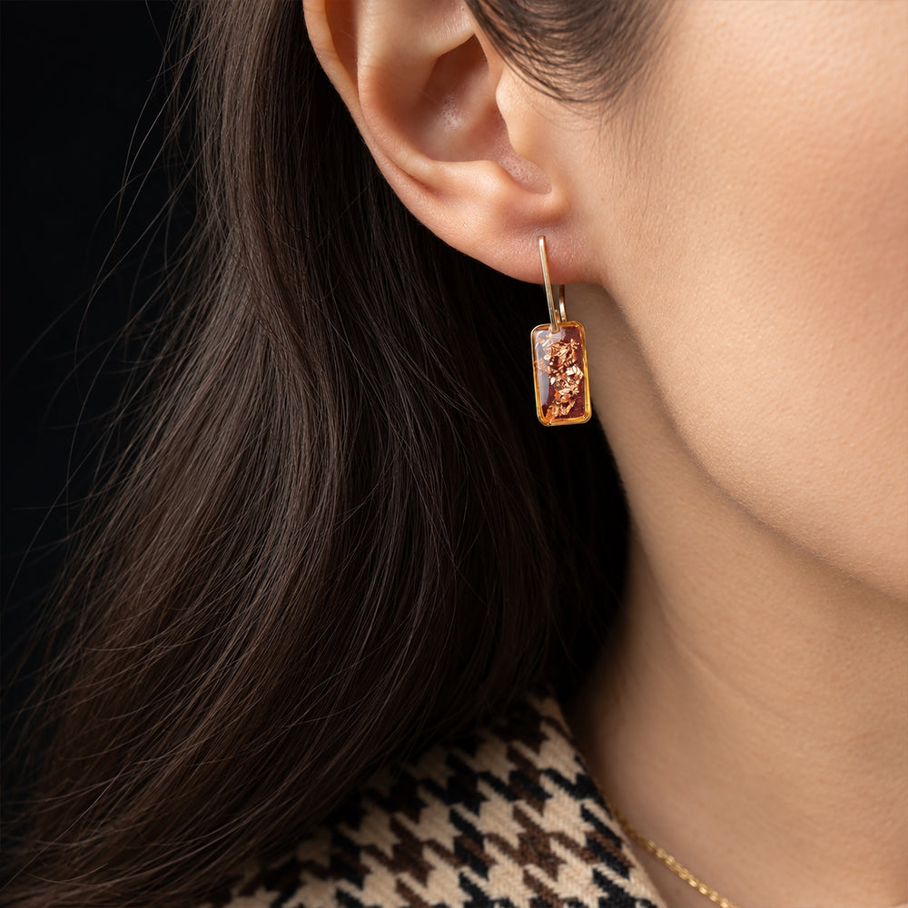 A pair of gold drop earrings featuring rectangular amber resin pendants with embedded copper-colored metallic flecks, set against a clean off-white background.