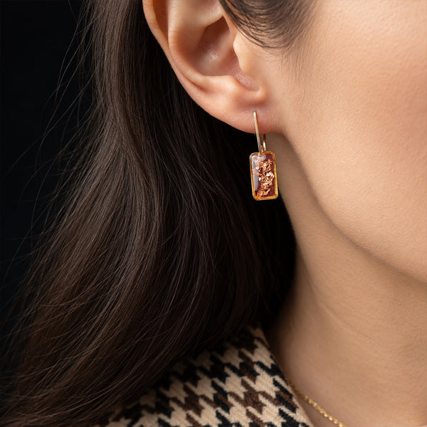 A close-up of a woman's ear wearing a small rectangular gold-toned earring with a raised classical figure design, shown against dark hair and a black background.