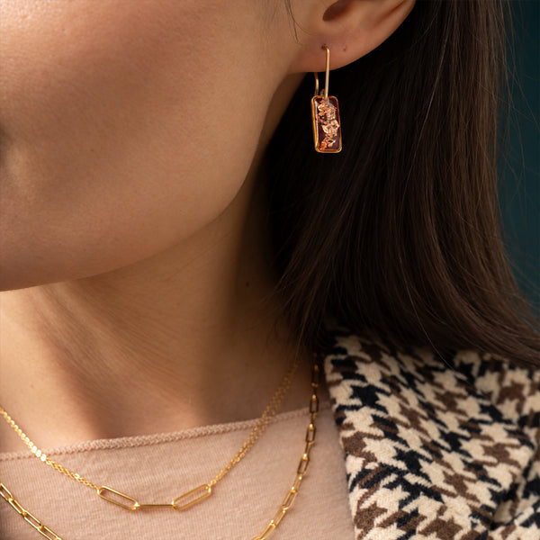 A close-up lifestyle shot of a model wearing a gold rectangular drop earring with a textured, organic gold pattern inside. The model is also wearing layered gold necklaces and a brown, black, and beige houndstooth patterned jacket against a dark teal background.
