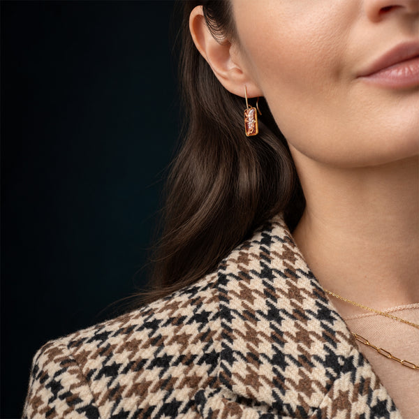 A close-up lifestyle shot of a woman wearing a delicate gold dangle earring with a rectangular amber stone, paired with a brown and beige houndstooth blazer.