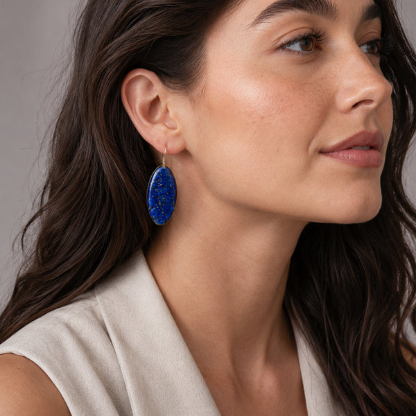 A close-up of a woman wearing a oval blue stone drop earring with tiny gold flecks, paired with a light beige top against a soft gray background.